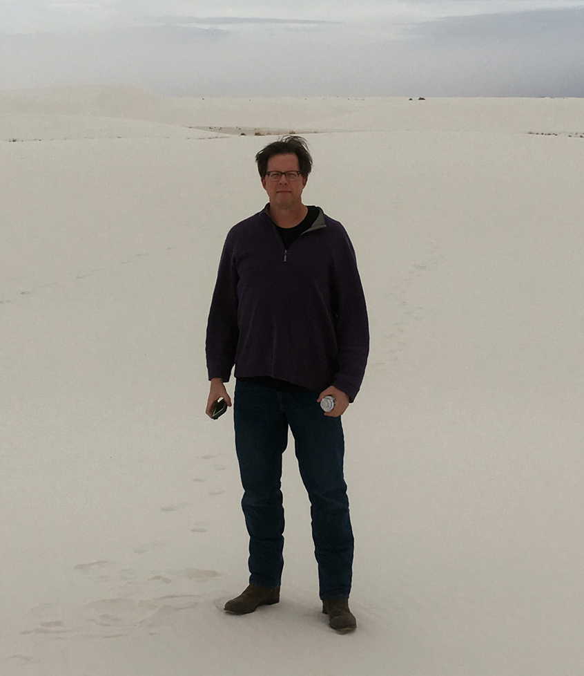 Joe Forman stands atop a sand dune, wearing a dark fleece jacket and jeans. In one hand he is holding a phone, in the other he is carrying a drink. The sky is overcast.